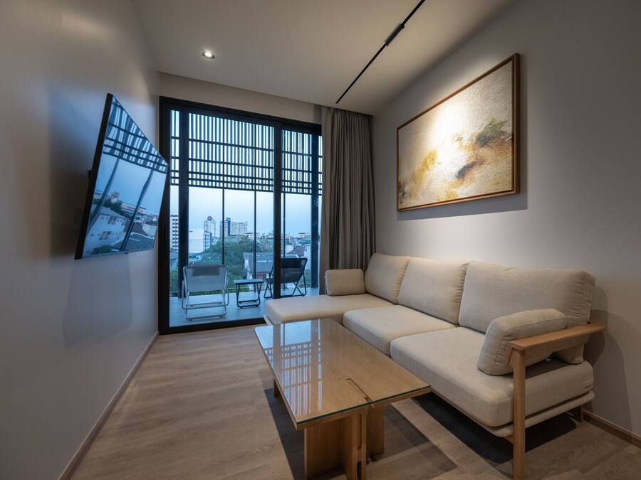 A modern living room with a sofa, a glass coffee table, a TV mounted on the wall, and a large window with a view of buildings.