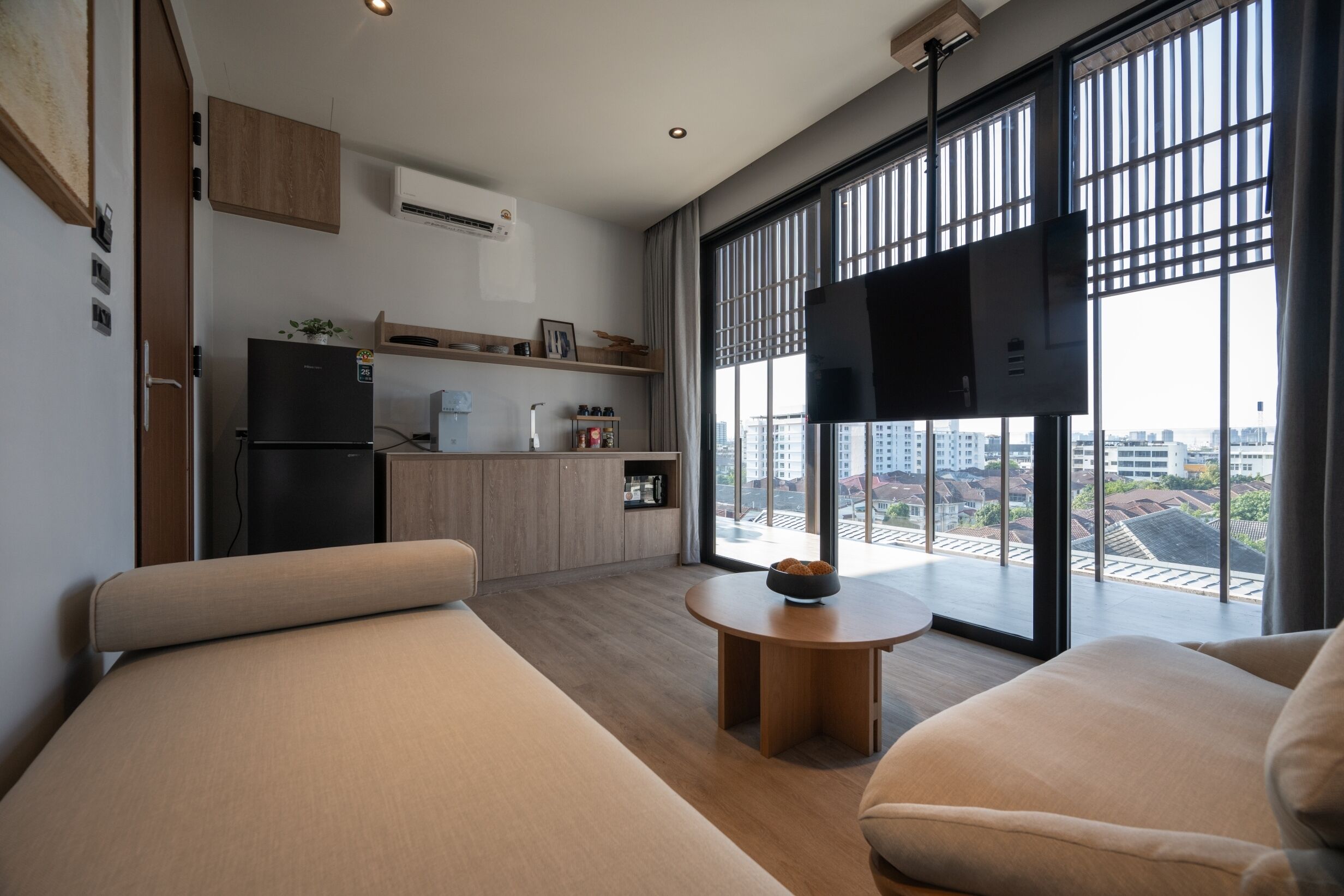 A modern living room with a sofa, a small round table, and a kitchen area with a refrigerator and a flat-screen TV.