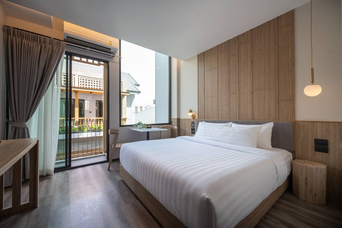 Ett modernt hotellrum med en stor säng, ett skrivbord och en balkong med utsikt över byggnaderna.