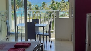 A room with a bed, a table, and chairs, a sliding glass door leading to a balcony with a view of the ocean and palm trees. - Ocean view, 2-minute walk to the beach (Le Moule)