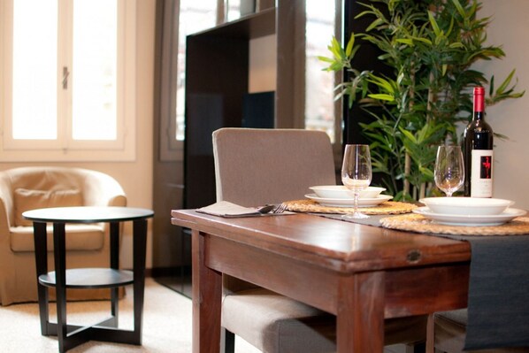 A dining area with a wooden table set for two, a small round table, a sofa, and a potted plant.