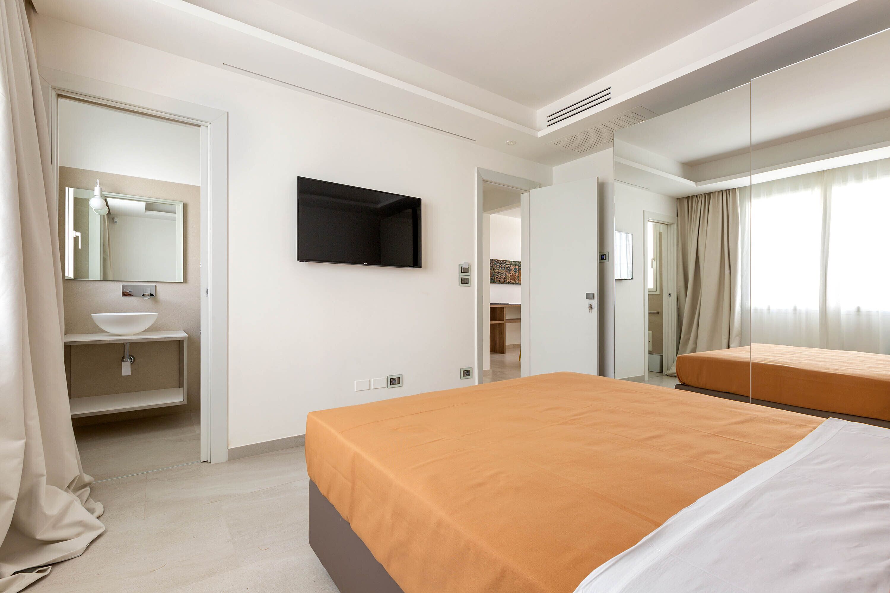 Camera d'albergo con un letto grande, un comodino, uno specchio, una TV a schermo piatto e un bagno con lavandino e specchio.