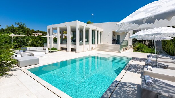 Un espace moderne aménagé au bord de la piscine, avec une piscine à l’eau d’un bleu limpide, des meubles d’extérieur blancs et un bâtiment contemporain doté de grandes fenêtres et d’un patio couvert.