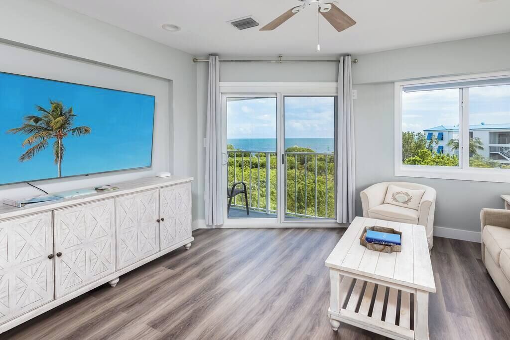 A spacious living room with a large TV, a white coffee table, and a balcony with a view of the ocean.