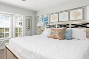 Un dormitorio con una cama grande, ropa de cama blanca y un cabecero con diseño de travesaño. Hay tres obras de arte enmarcadas sobre la cama, y una ventana con persianas a la izquierda.