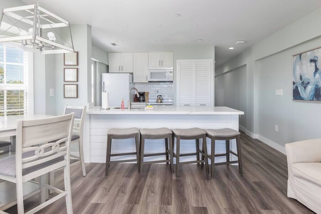 Una cocina moderna con armarios blancos, una isla central con taburetes y un comedor con mesa y sillas blancas.
