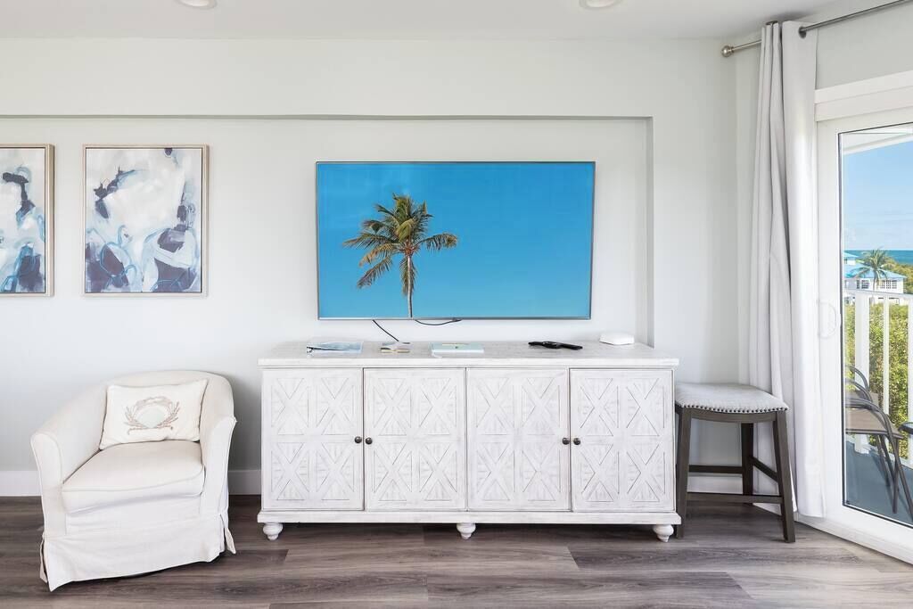A room with a white cabinet, a white armchair, a small stool, and a painting of a palm tree.