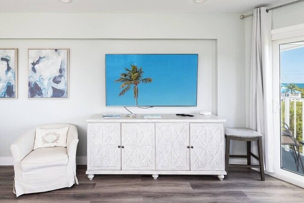 A room with a white cabinet, a white armchair, a small stool, and a painting of a palm tree.