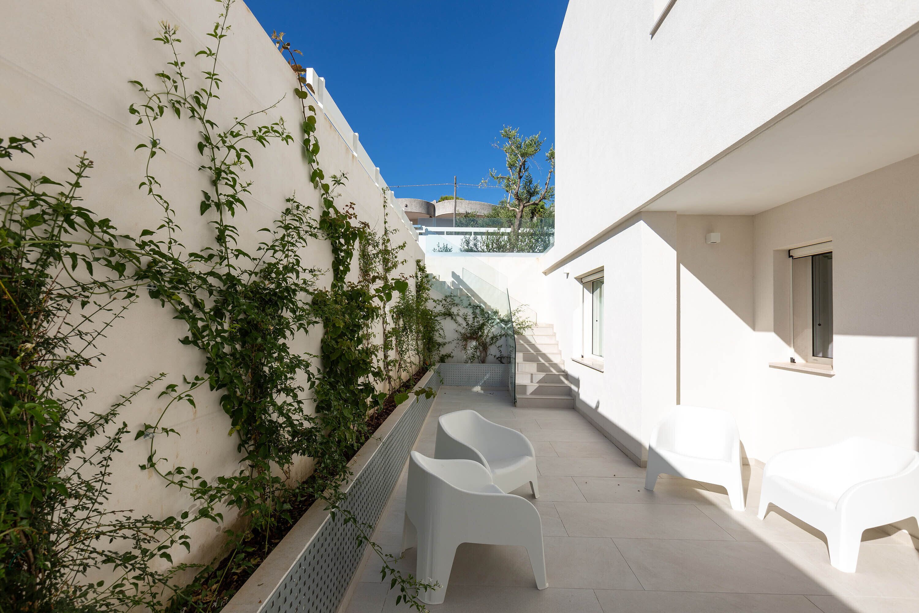 A narrow outdoor corridor with white walls, a small garden area with climbing plants, and two white plastic chairs.