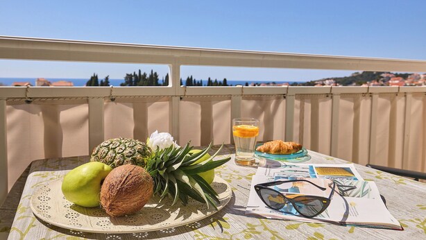 A table with fruits, a glass of juice, a croissant, and sunglasses on a balcony overlooking a coastal view. - Apartment Terra - Three Bedroom Apartment, Sleeps 5 (Dubrovnik)