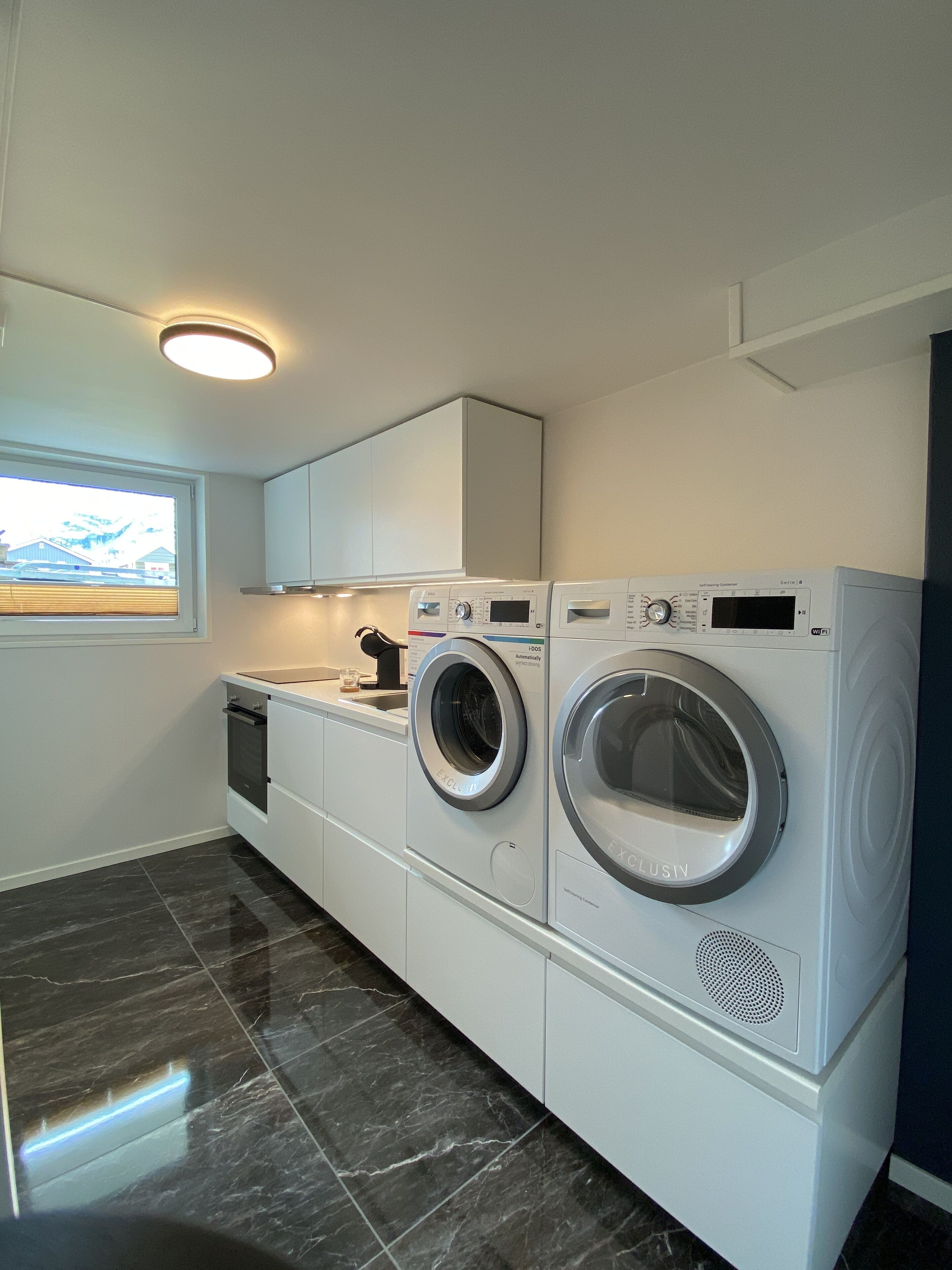 A modern laundry room with two front-loading washing machines, a built-in oven, and a window with a view of a mountain.