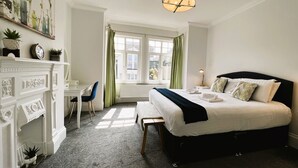 Une chambre avec un grand lit, un bureau et une chaise. Il y a une fenêtre avec des rideaux, un lustre et un radiateur.