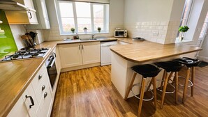 Une cuisine compacte équipée d’une cuisinière à gaz, d’un four à micro-ondes et d’un évier. On y trouve des tabourets en bois au comptoir et une fenêtre agrémentée de plantes en pot.