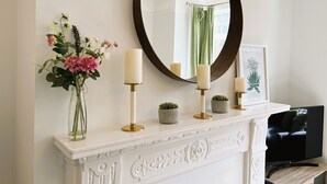 Une cheminée blanche ornée de trois bougies, un vase de fleurs et une illustration botanique encadrée.