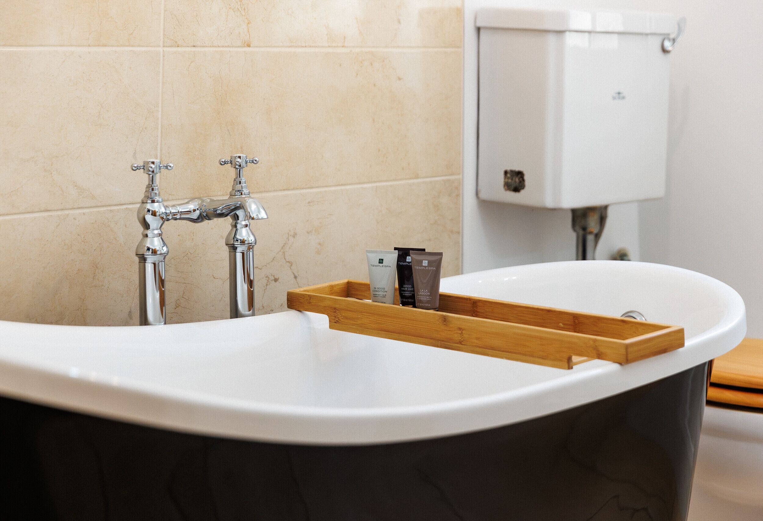 A bathroom with a white sink, two chrome faucets, a wooden tray, and a toilet tank.