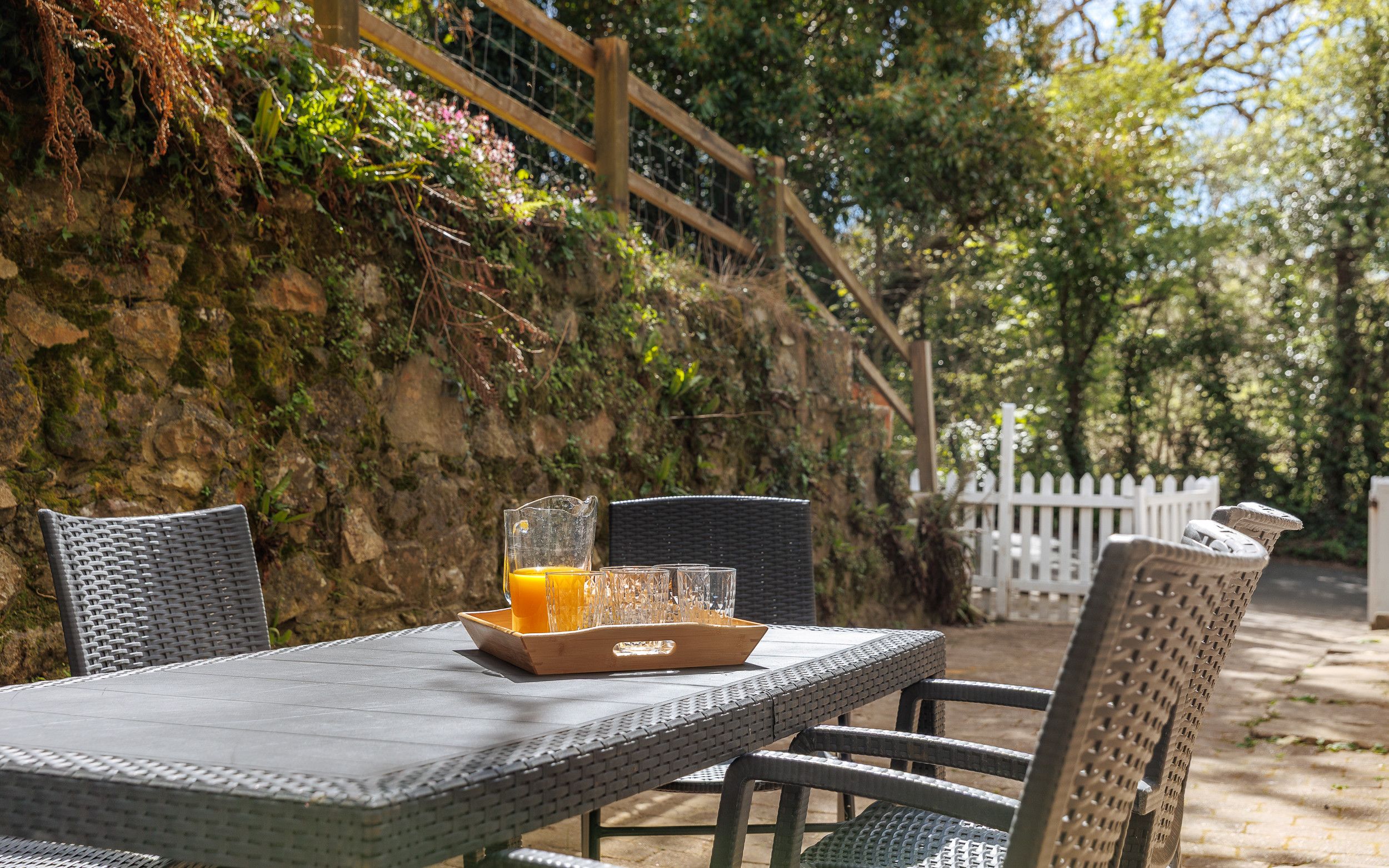 Une table d’extérieur avec des chaises en osier, un plateau avec un verre de jus d’orange, et une clôture blanche en bois en arrière-plan.