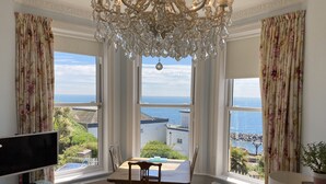 Ein Zimmer mit Esstisch, Stühlen, Fernseher und einer Chandelier. Große Fenster bieten Blick auf das Meer und Gebäude.