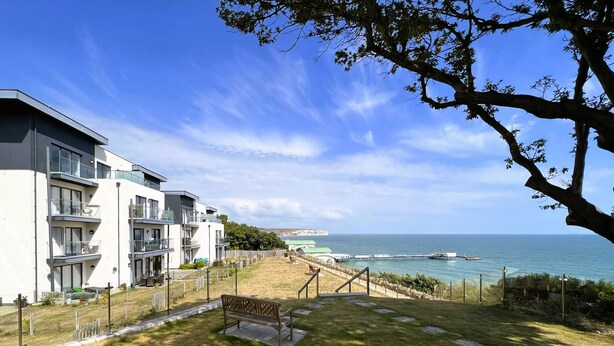 A residential area with a grassy lawn, a bench, and a view of the sea. - New Horizon (Sandown)