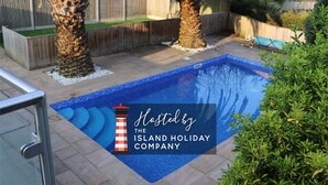 A swimming pool with a tiled deck, surrounded by palm trees and a wooden fence. - No 1 Ashey (Ryde)