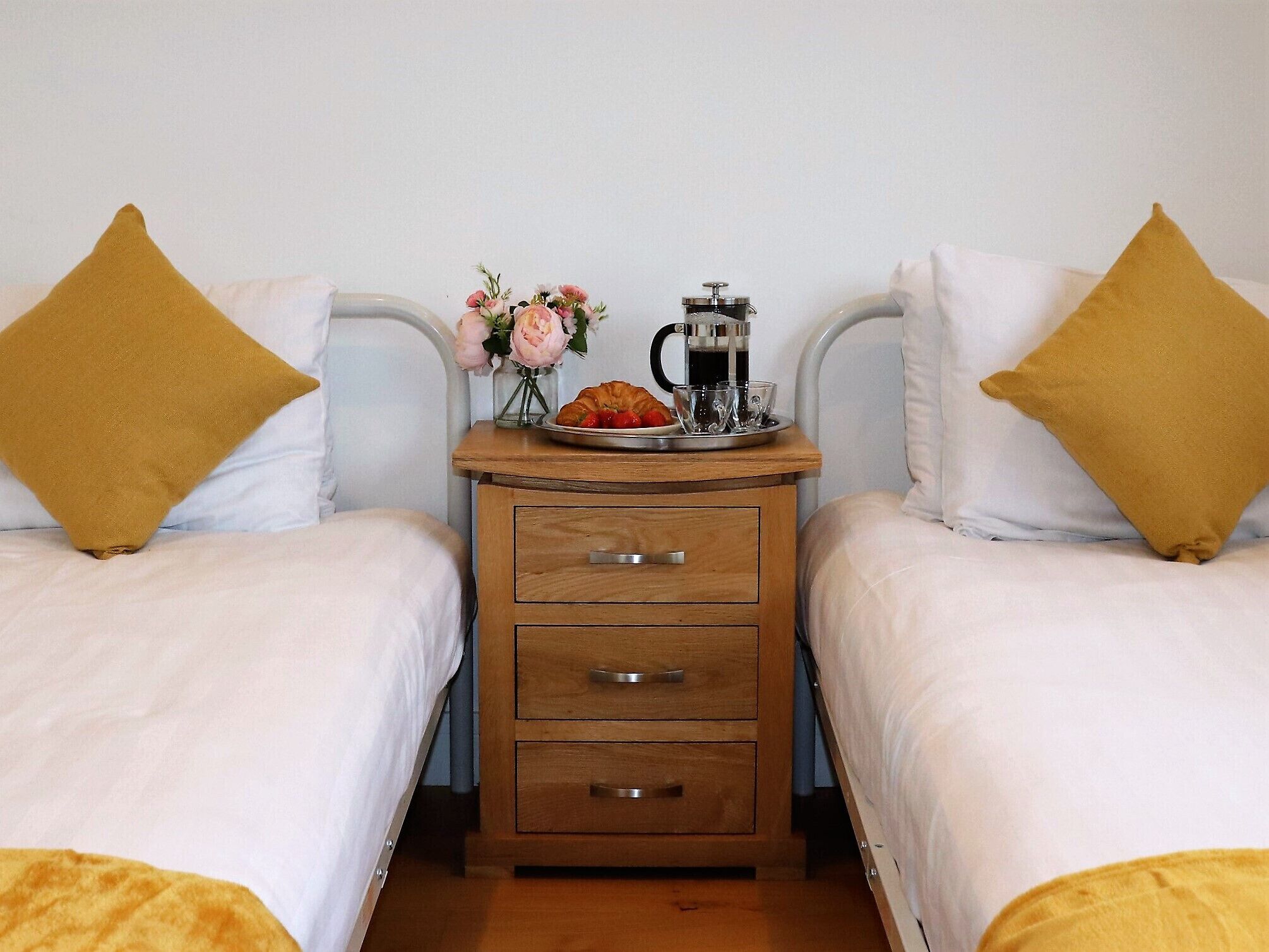 Een hotelkamer met twee eenpersoonsbedden, een houten nachtkastje met een koffiezetapparaat, een bord croissants en een bloevas.