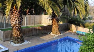 A swimming pool with a tiled deck, surrounded by palm trees and a wooden fence.