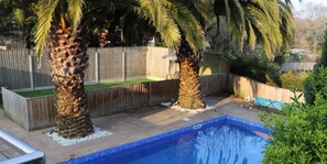 A swimming pool with a tiled deck, surrounded by palm trees and a wooden fence. - No 1 Ashey (Ryde)