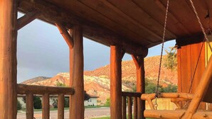 A wooden porch with a swing, a dog sitting on the deck, and a view of red rock hills in the background. - The Zen Den (Cedar City)