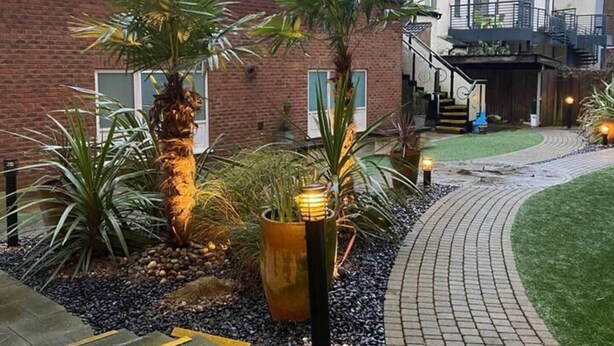 A landscaped garden area with a brick building, palm trees, and a pathway leading to a staircase.
