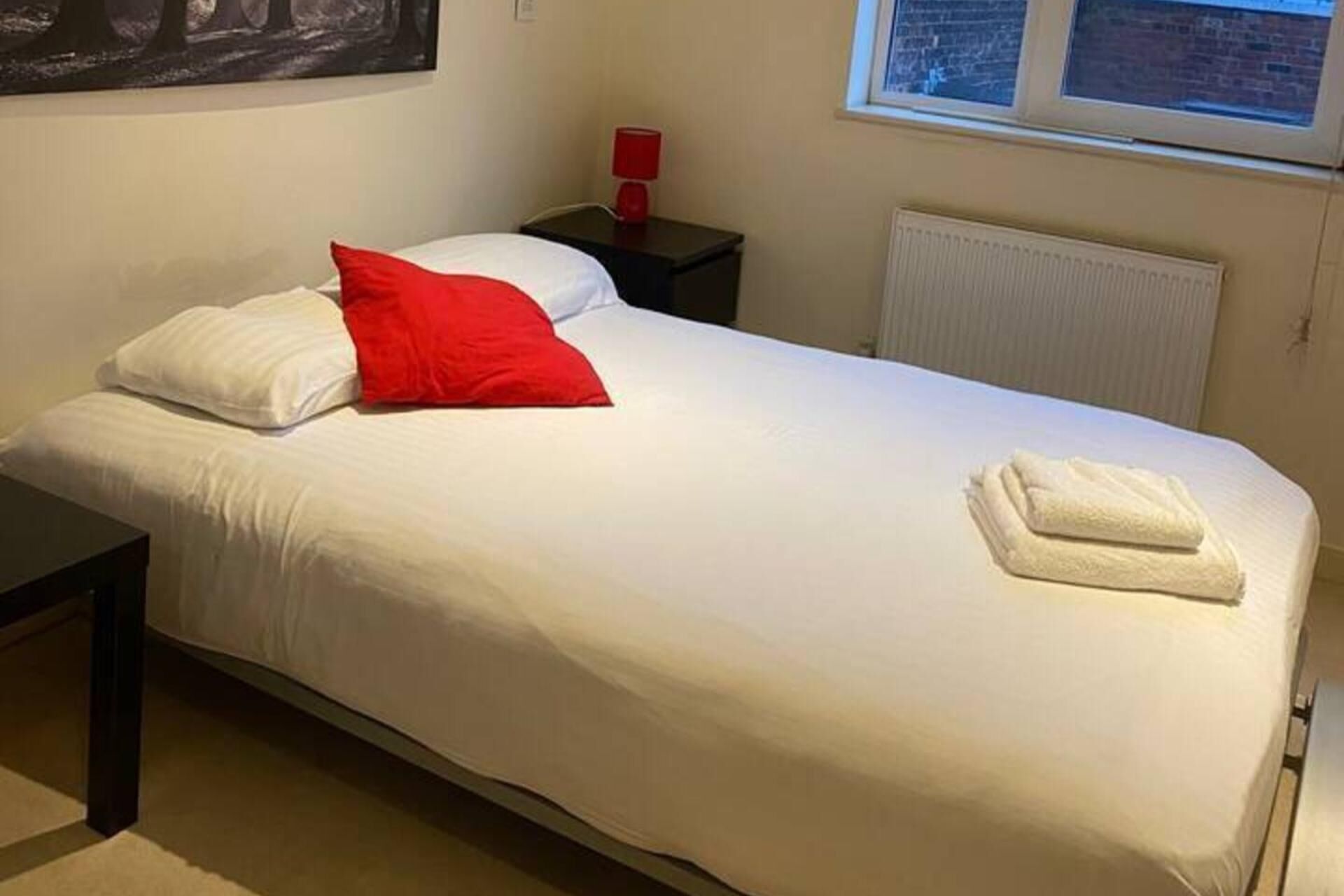 A single bed with white bedding and a red pillow, a small bedside table with a red lamp, and a window with a view of the outside.