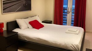 A neatly made bed with white linens and a red pillow, a small bedside table with a red lamp, and a window with red curtains.