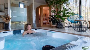 Une femme se détend dans un bain à remous dans une salle de bain moderne, avec une grande fenêtre, une plante en pot et une chaise bleue.