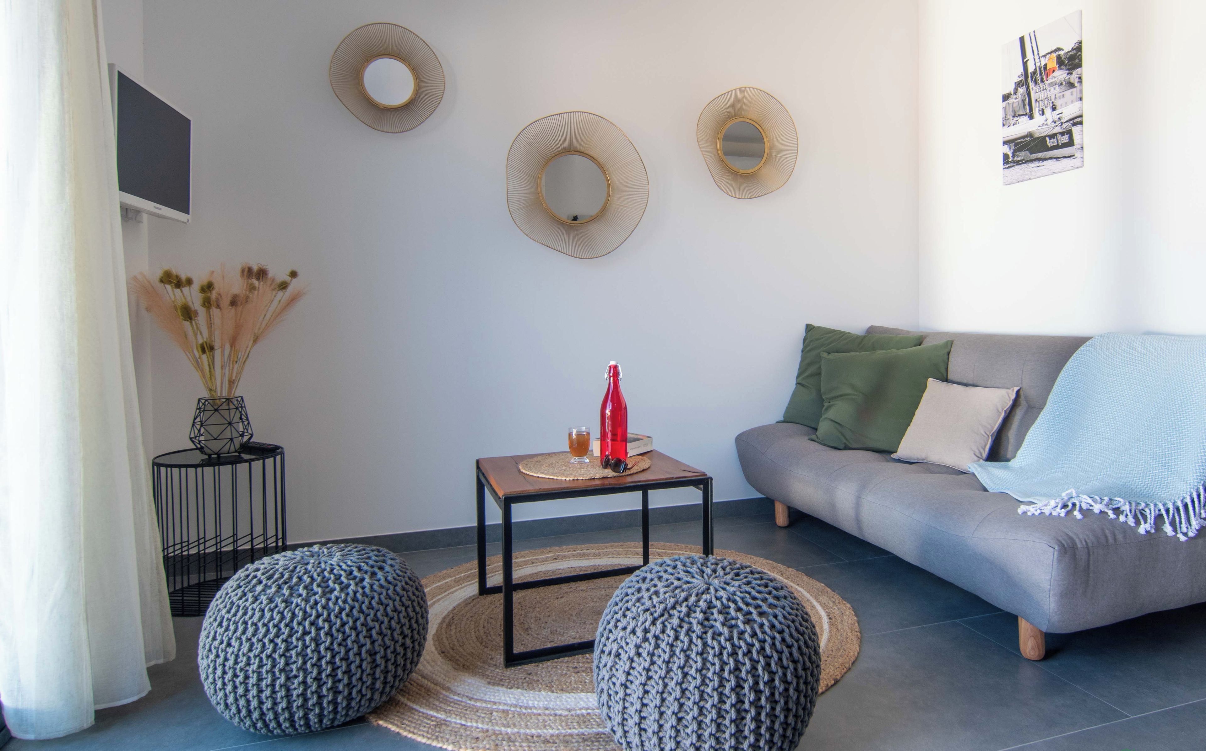 Ein modernes Wohnzimmer mit einer grauen Couch, zwei runden Hocker, einem kleinen Holztisch mit einer roten Vase und einem gewebten Teppich.