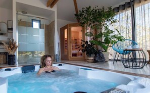 Une femme se détend dans un bain à remous dans une salle de bain moderne, avec une grande fenêtre, une plante en pot et une chaise bleue.
