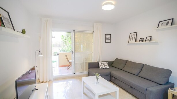 Sala de estar moderna com sofá cinzento, mesa de centro branca e um abajur. Há uma porta de vidro deslizante que dá para uma área externa com uma cadeira e uma mesa.