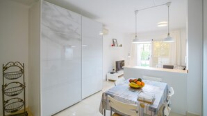 Uma cozinha moderna com armários brancos, uma mesa de jantar com uma fruteira e uma janela com vista para o verde.