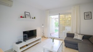Uma sala de estar moderna com uma TV de tela plana sobre um suporte branco, um sofá cinza com almofada de bolinhas, uma mesa de centro branca e uma porta de vidro deslizante que dá para uma área externa.