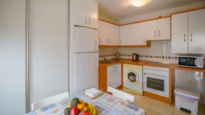 Cozinha compacta com armários brancos, máquina de lavar, micro-ondas e uma mesa com frutas.