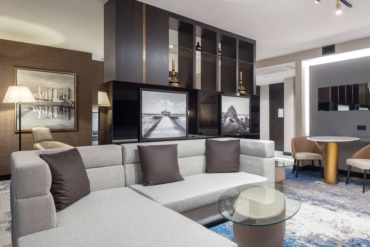 Une chambre d’hôtel moderne avec un canapé gris, une table basse en verre, une table à manger ronde avec des chaises et des œuvres d’art accrochées aux murs.