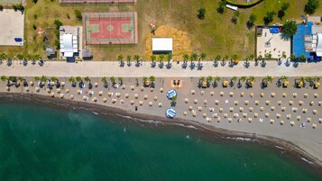 Vista aérea de una playa con filas de tumbonas y sombrillas, una cancha de tenis y un edificio con techo azul.