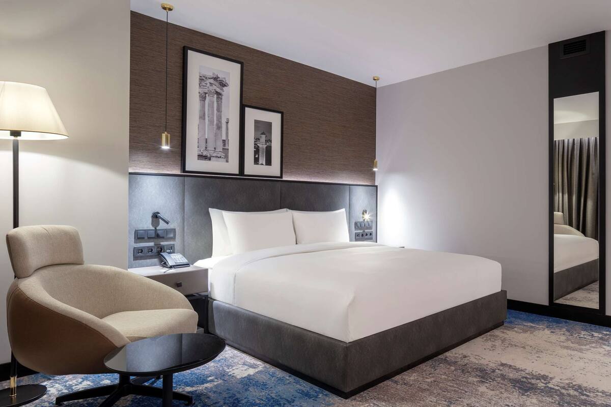 Une chambre d’hôtel moderne, dotée d’un grand lit, d’une chaise, d’un bureau, d’une lampe et de tableaux encadrés au mur.