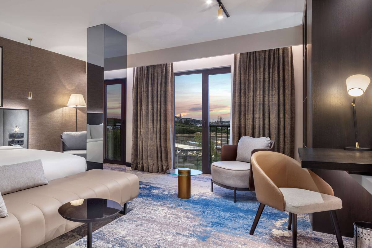 Une chambre d’hôtel moderne, dotée d’un grand lit, d’un canapé, d’un fauteuil, d’une petite table et offrant une vue sur la ville grâce à de grandes fenêtres.