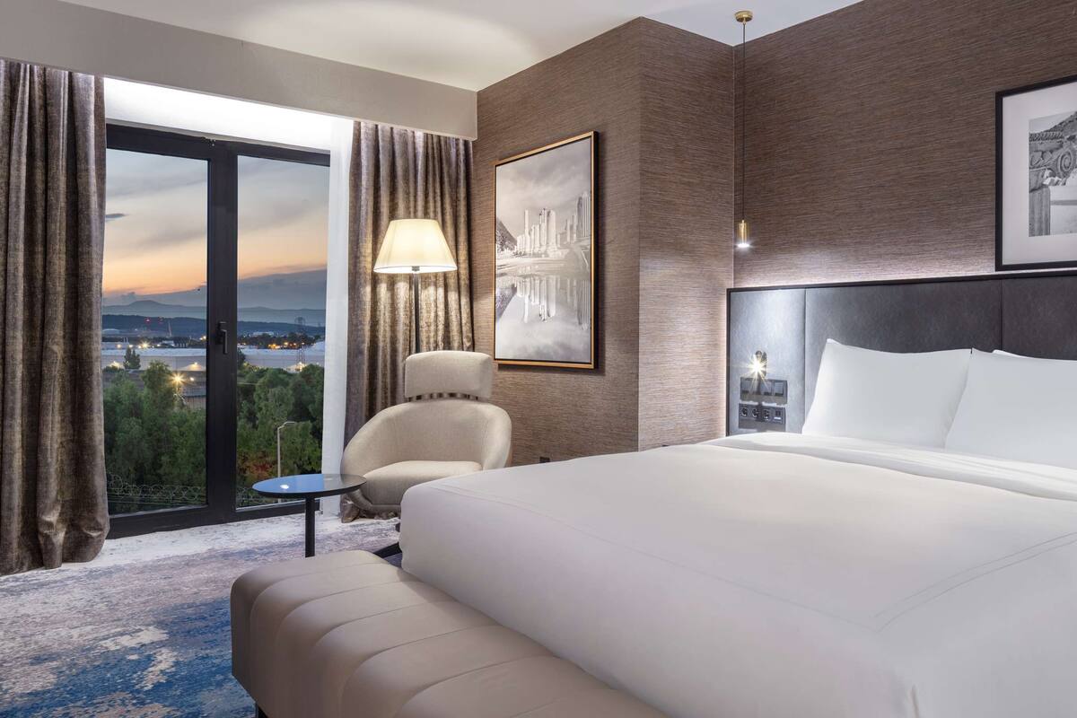 Une chambre d’hôtel avec un grand lit, une chaise, une petite table et une vue sur la ville grâce à de grandes fenêtres.