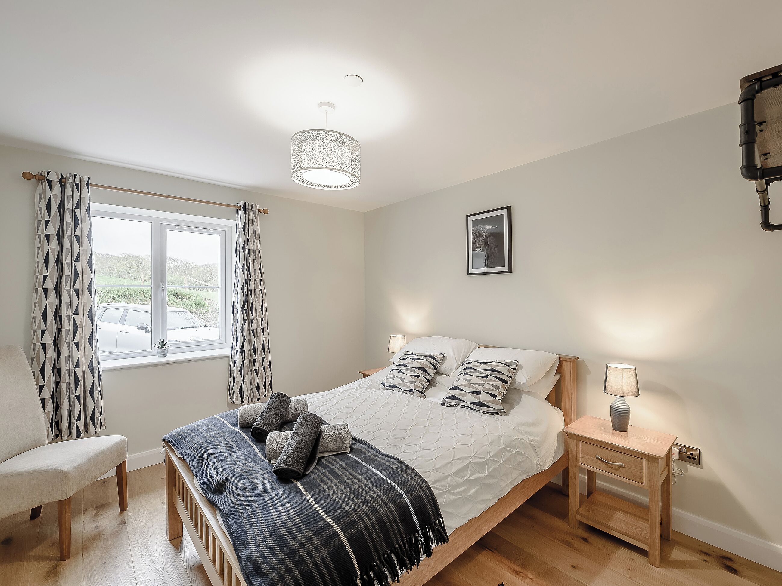 Een slaapkamer met een bed, nachtkastjes, een stoel, een raam met gordijnen en een plafondlamp.
