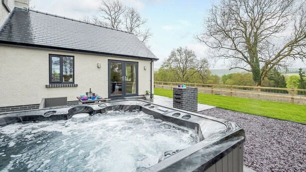 Un espace de détente avec un jacuzzi, une petite table sur laquelle sont disposés divers objets, et une vue sur une maison et le paysage environnant.