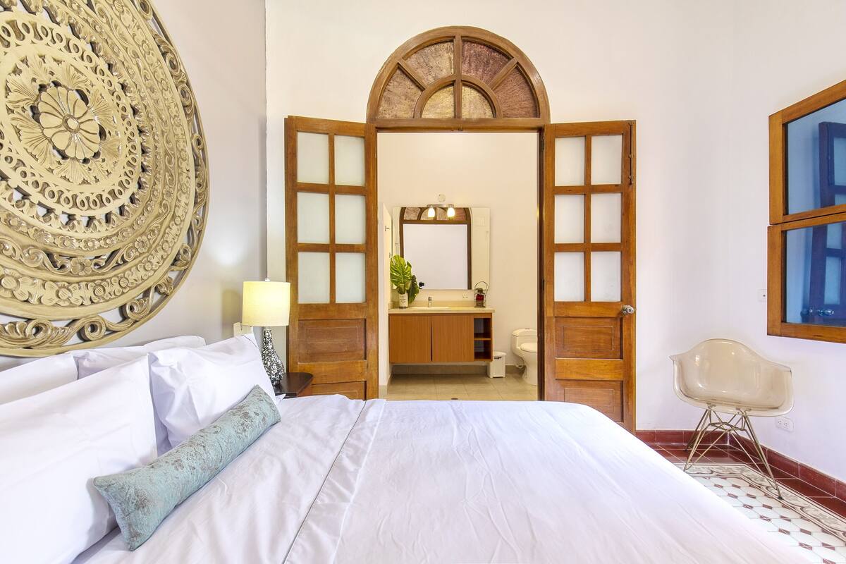 A hotel room with a large bed, a wooden door leading to a bathroom, and a decorative wall piece.