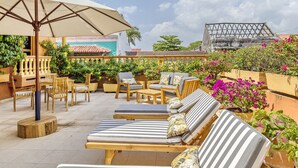 A rooftop terrace with lounge chairs and umbrellas, surrounded by greenery and a building with a yellow facade.