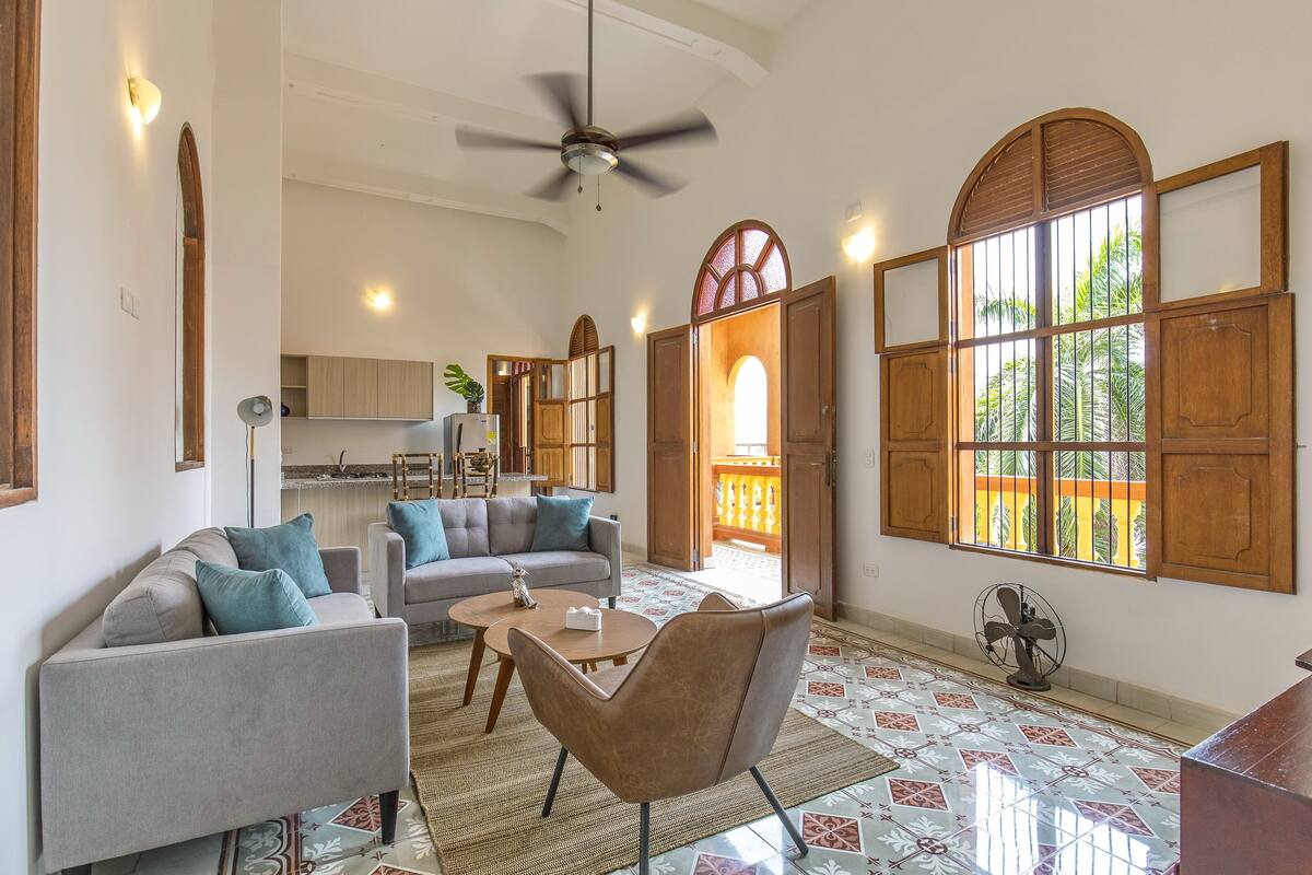 A spacious living area with a sofa, armchair, and coffee table, a ceiling fan, and large windows with wooden shutters.