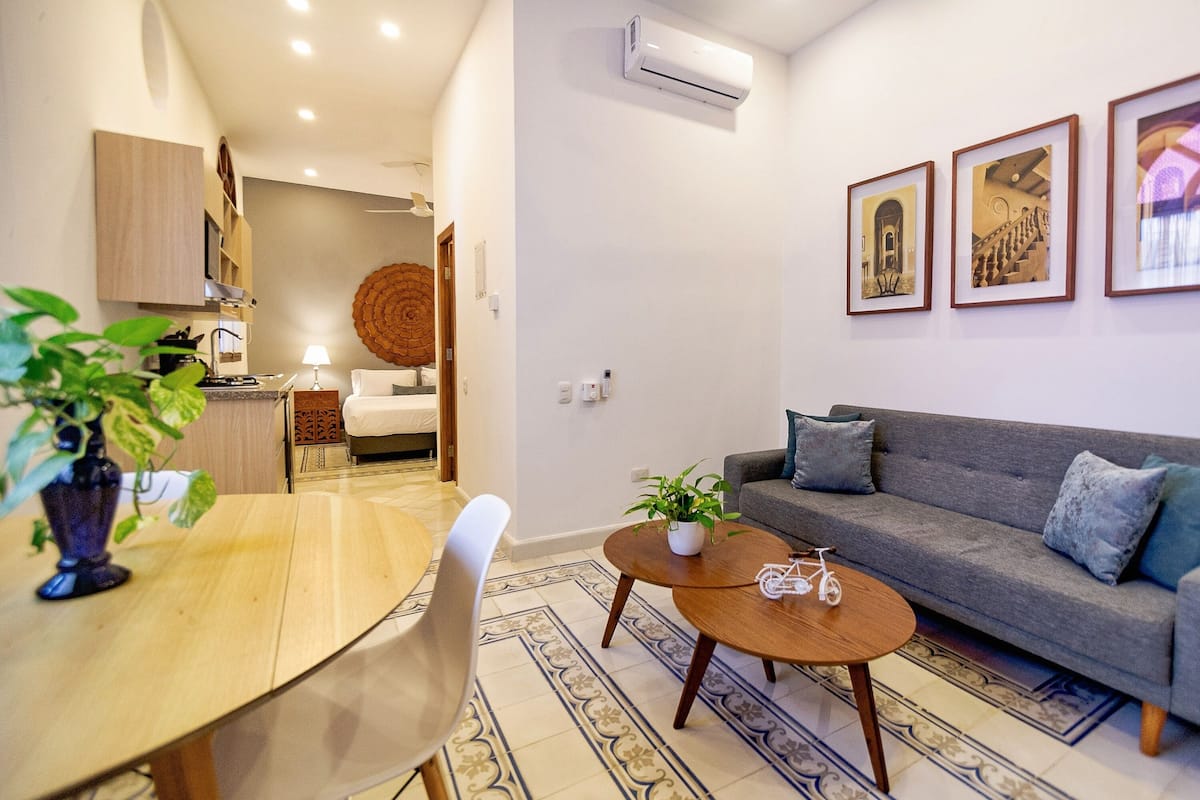 A modern living room with a grey sofa, a wooden coffee table, and a dining area with a table and chairs. There are framed artworks on the wall and a ceiling fan above the sofa.