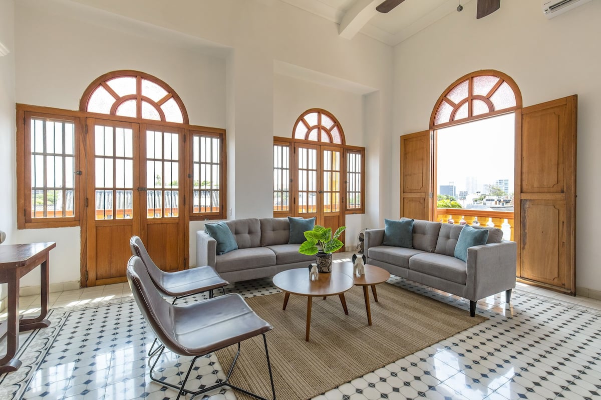 A spacious living room with a grey sofa, a coffee table, and a modern chair. Large wooden-framed windows allow natural light to fill the room.