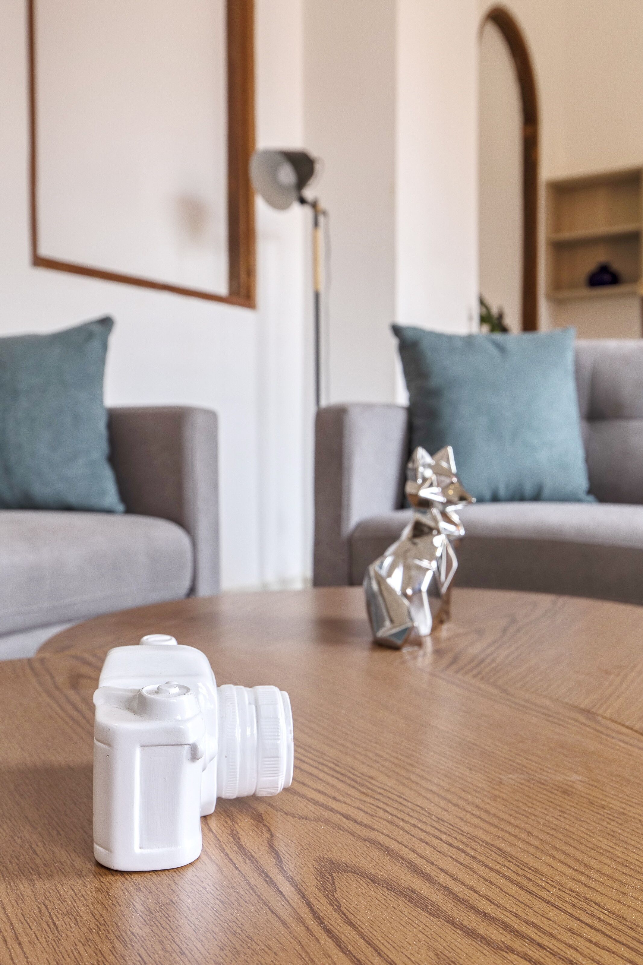 Ein Holztisch mit zwei weißen Flaschen und einer Metallskulptur in einem Wohnzimmer mit grauen Sofas und türkisfarbenen Kissen.